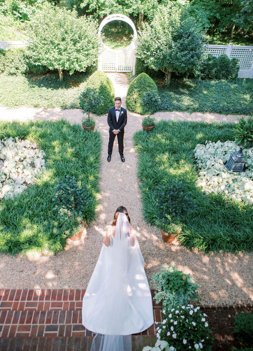 4th of July Wedding: Aerial view of a bride walking towards her groom along a pebbled garden path lined with green and white foliage.