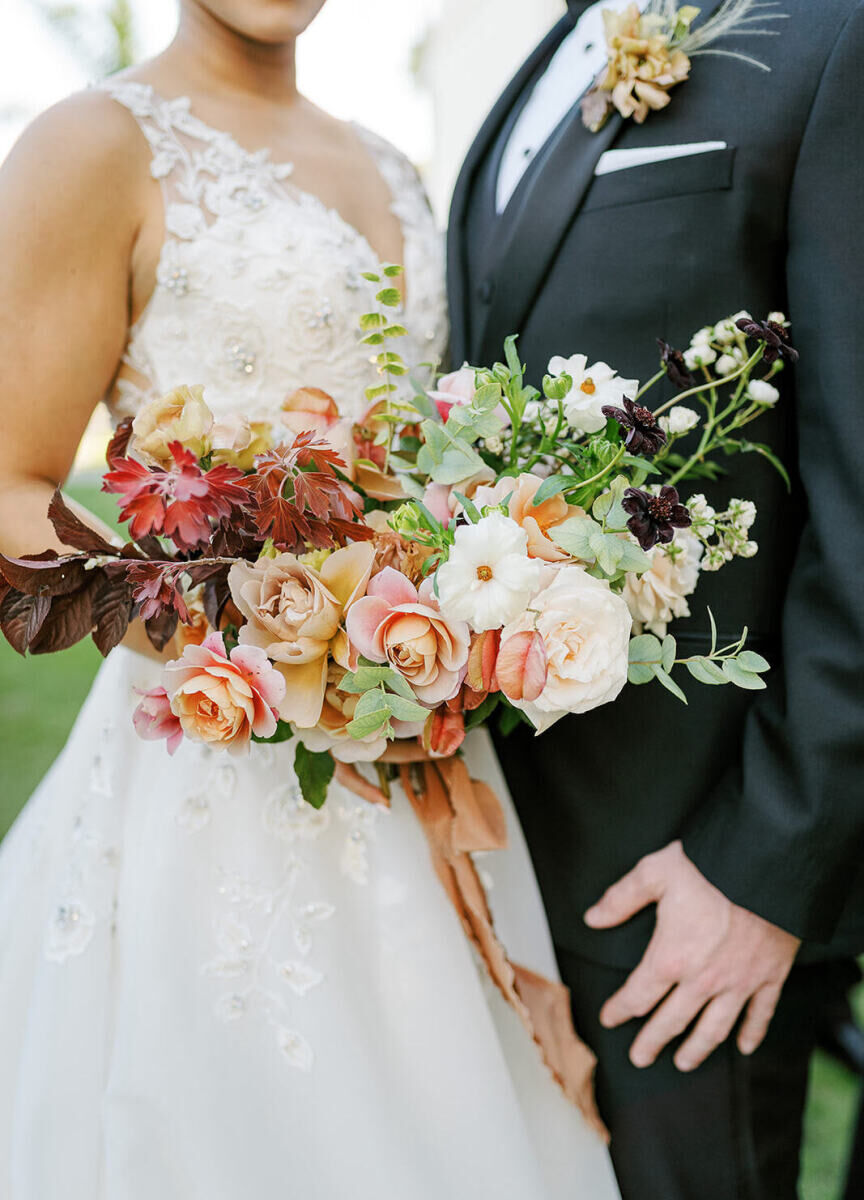 A bride holds her earth tone wedding bouquet, which features roses, Japanese ranunculus, and assorted foliage in shades of brown, red, and green.