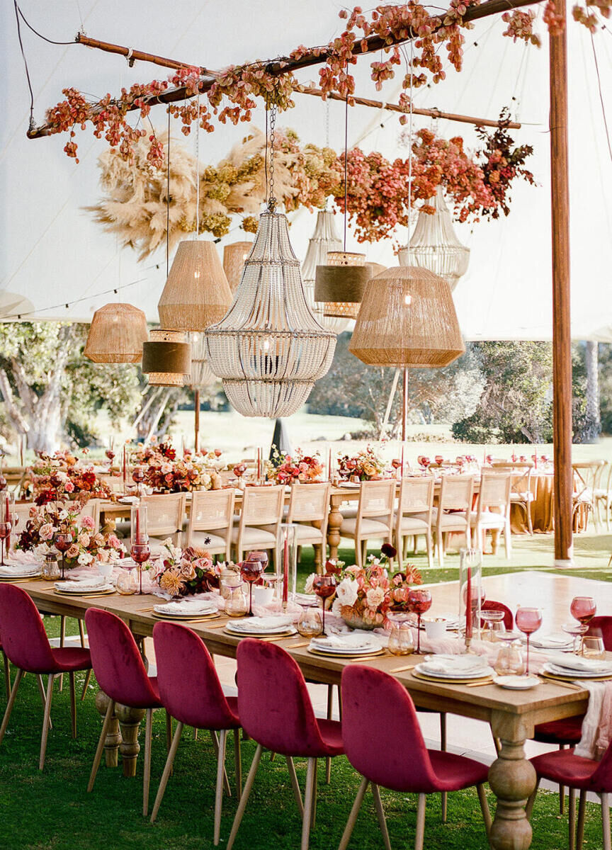 An earth tone wedding reception set with rich raspberry velvet chairs, a collection of hanging light fixtures, and an ombre floral installation starting in warm cream tones, progressing to yellows and corals, and ending in deep burgundy. 