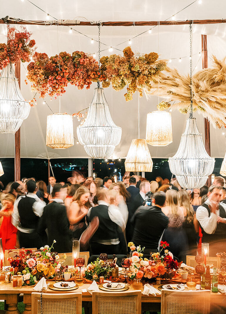 Guests fill the dance floor at an earth tone wedding reception under a tent filled with textured decor and interesting lighting.