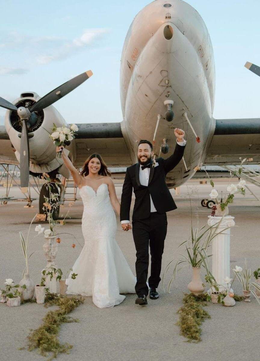 Adventurous Wedding Venues: A bride and groom standing in front of an airplane, holding hands with the other hands in the air, at Commemorative Air Force SoCal Hangar.