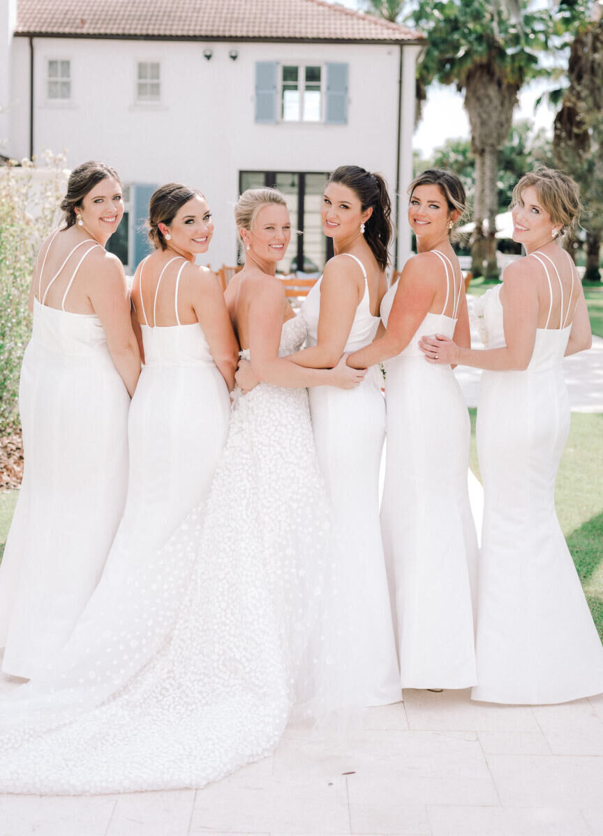 Bridesmaids in white surround the bride and smile for a photograph at an at-home wedding.