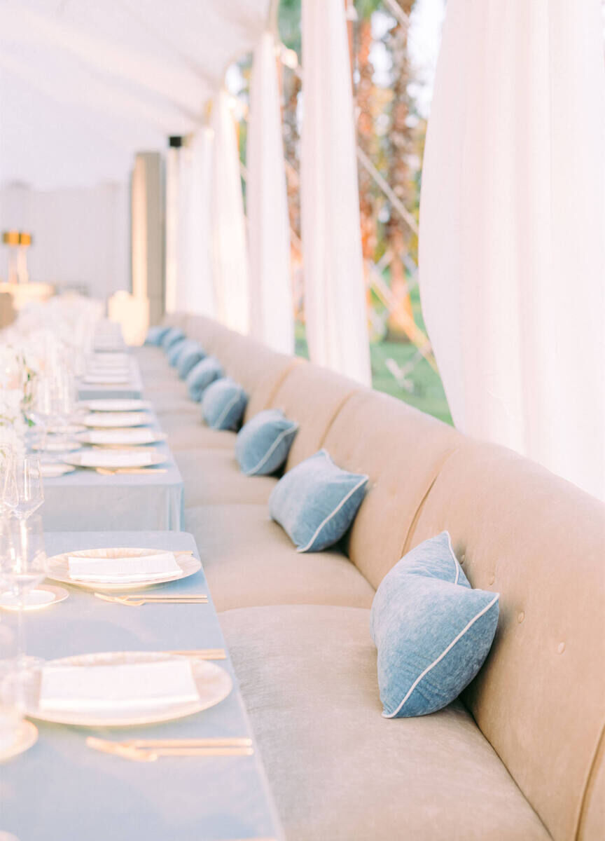 Beige banquettes set on the exterior sides of the long tables in a reception tent of an at-home wedding, with blue bolster pillows as accents.