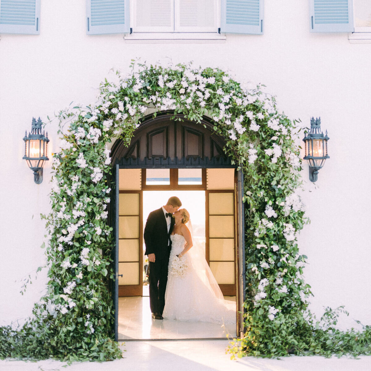 A bride and groom share a kiss inside the house where their at-home wedding took place, whose doorway was decorated with white flowers and greenery for the occasion.