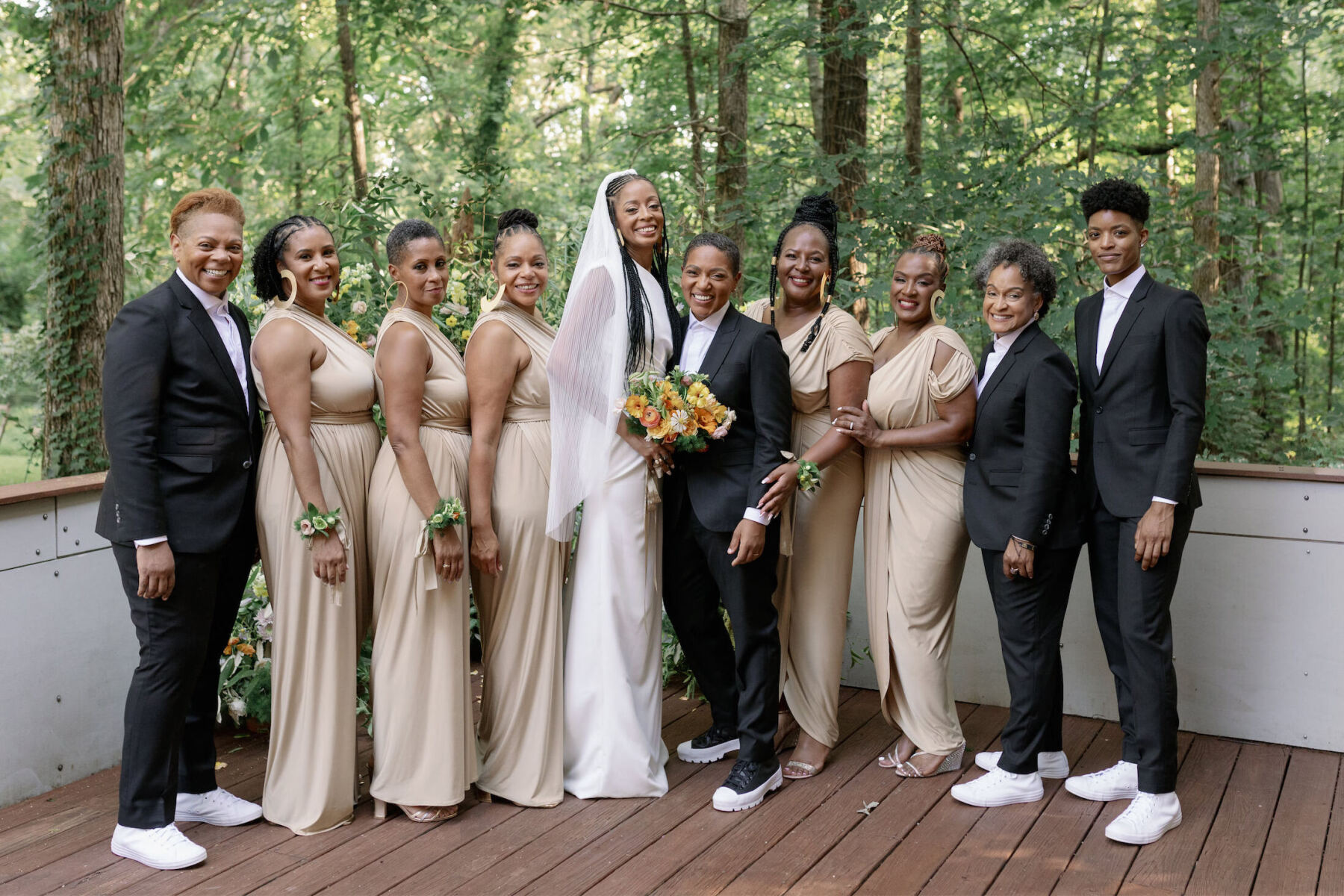 Two brides and their wedding party—in a mix of soft gold dresses and dark grey suits—are all smiles during a group portrait at their authentic modern wedding.