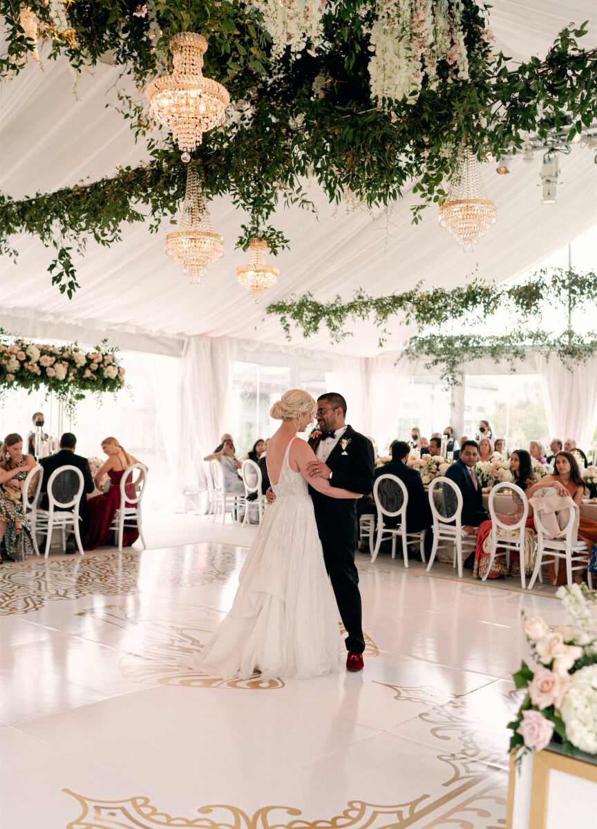 Wedding couple dancing at their tented wedding reception in front of guests featuring a greenery-forward hanging floral installation and crystal chandeliers 