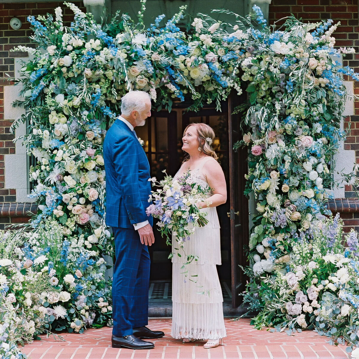A groom and bride look into each other's eyes in front of the pastel floral installation at the entrance to their home in St. Louis.