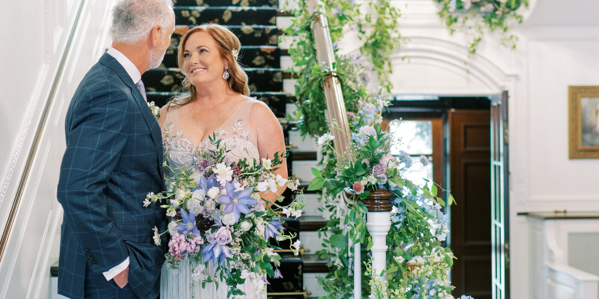 Wedding couple standing on a floral-adorned stairway, staring into each other's eyes.