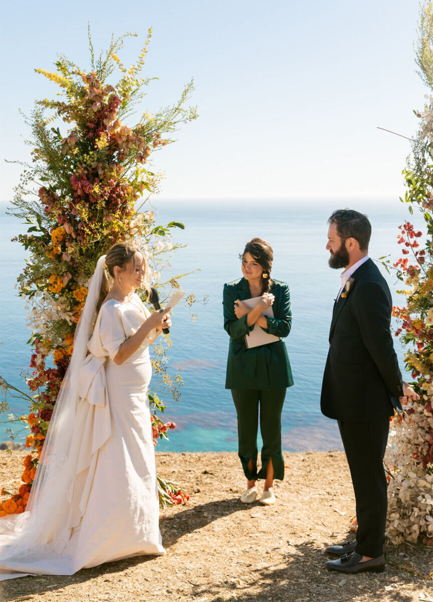 A bride recites her vows during her Big Sur wedding ceremony, with bold and colorful flowers behind her and the Pacific Ocean in the background.