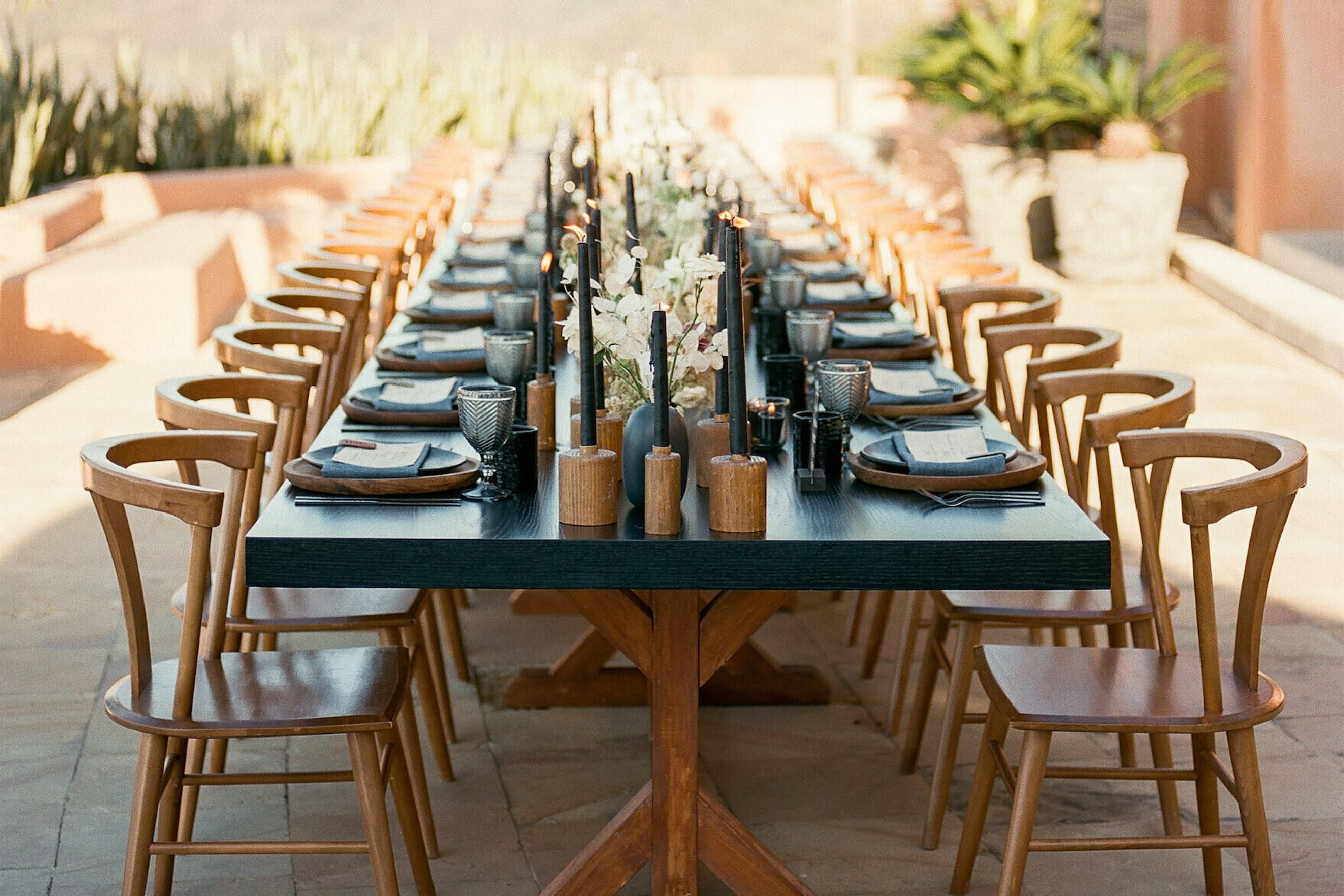 One long table for all of the guests at an intimate wedding reception, set with modern wood chairs, black taper candles, and handsome tabletop rentals.