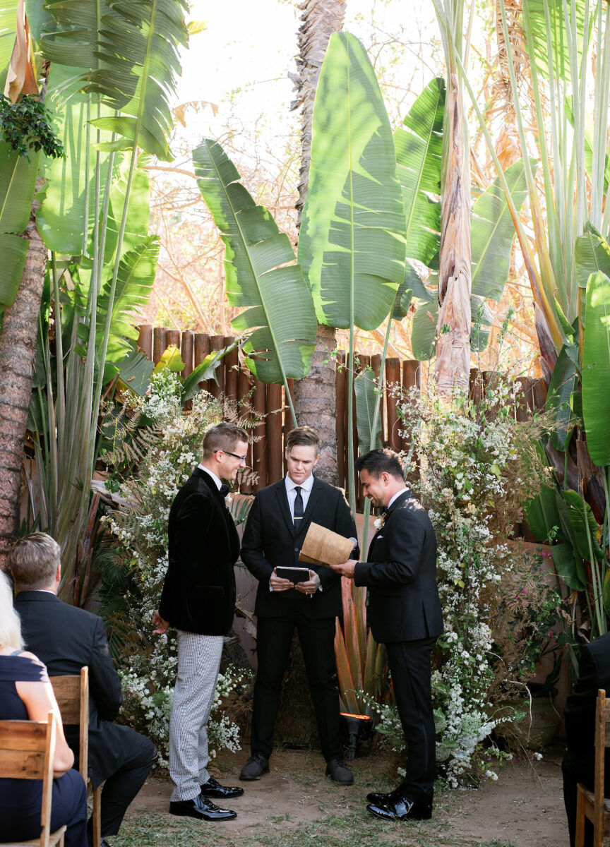A bohemian wedding ceremony setting, with a pair of tall floral pieces flanking the couple, who stood in front of tropical foliage.