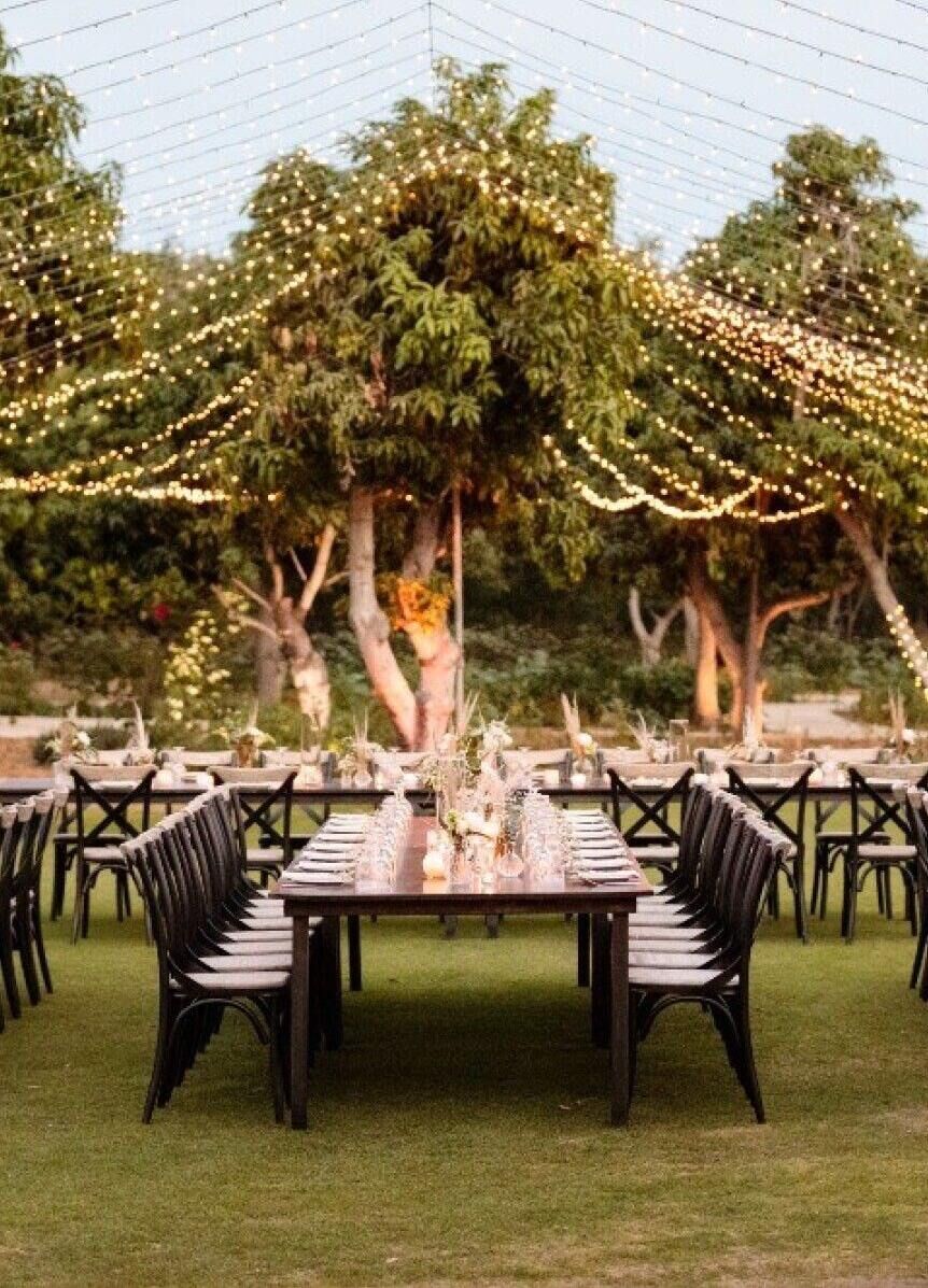Ceiling Wedding Decor: String lights hanging over an outdoor reception area with rectangular wooden tables.