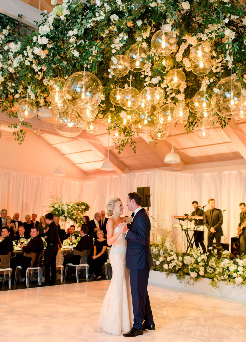 Ceiling Wedding Decor: A wedding couple dancing under an installation with greenery and white florals with large light bulbs.