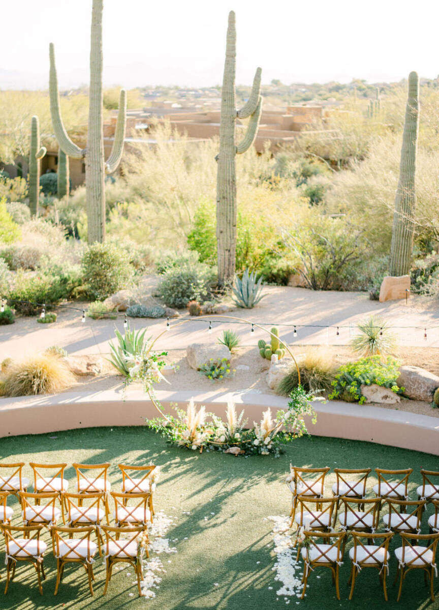 City Weddings: An outdoor wedding ceremony setup with wooden chairs and white seat cushions, a metal circular archway at the altar and cacti and the Arizona desert beyond.