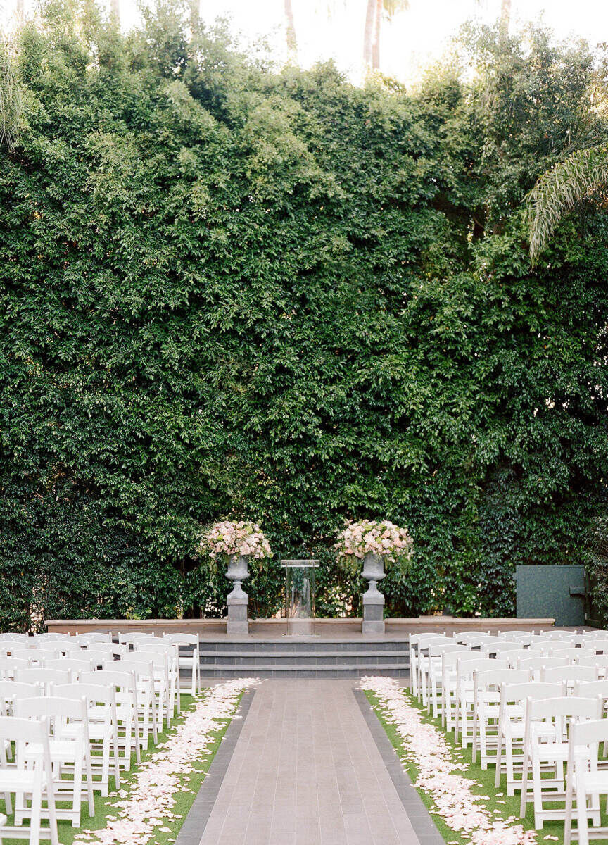 City Weddings: An outdoor wedding ceremony setup with white flower petals lining the aisle, white chairs for the guests, two floral arrangements and a wall of greenery.