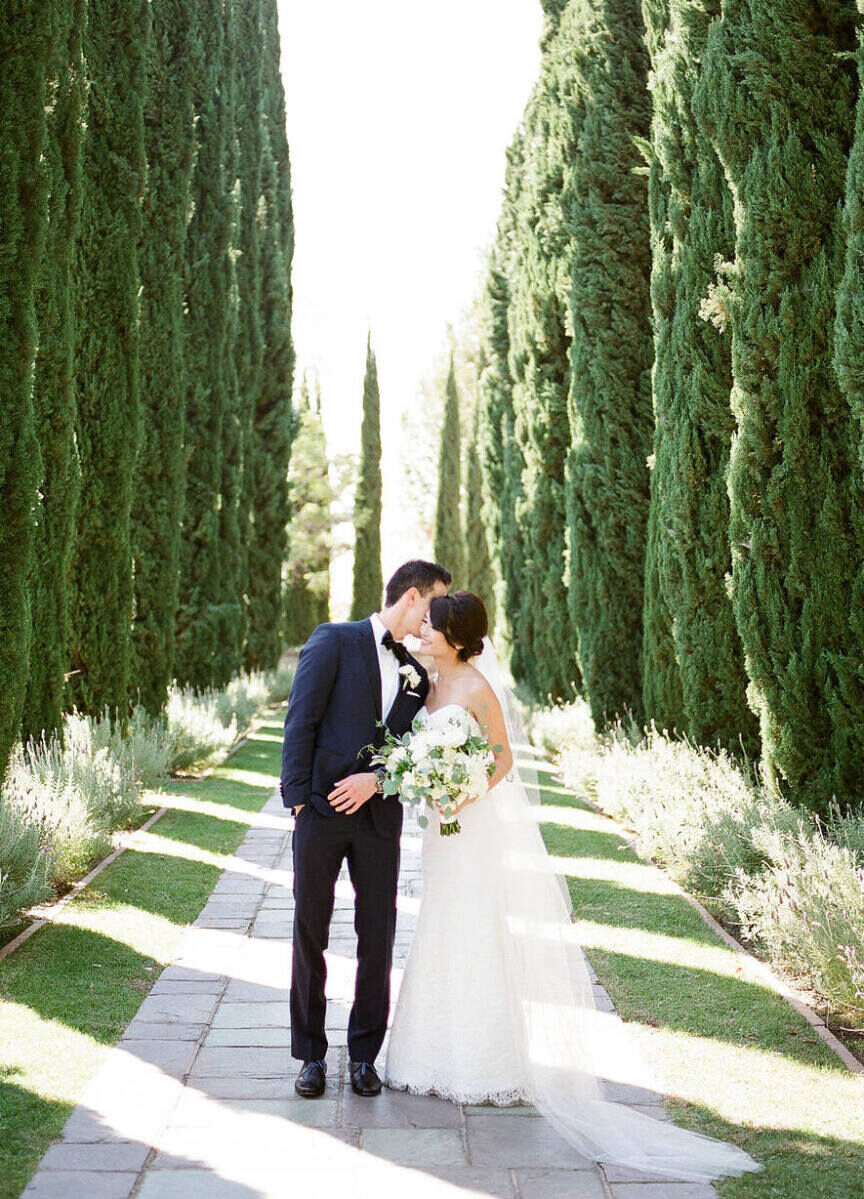 City Weddings: A groom kissing a bride while standing on a pathway lined with juniper shrubs.