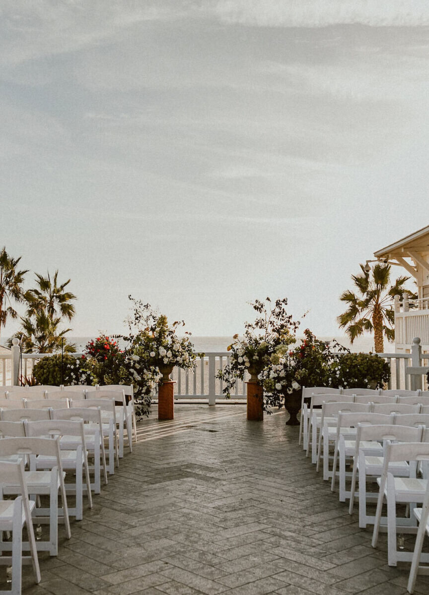 City Weddings: A wedding ceremony setup overlooking the ocean, with white chairs and two floral arrangements in oversize vases at the altar.