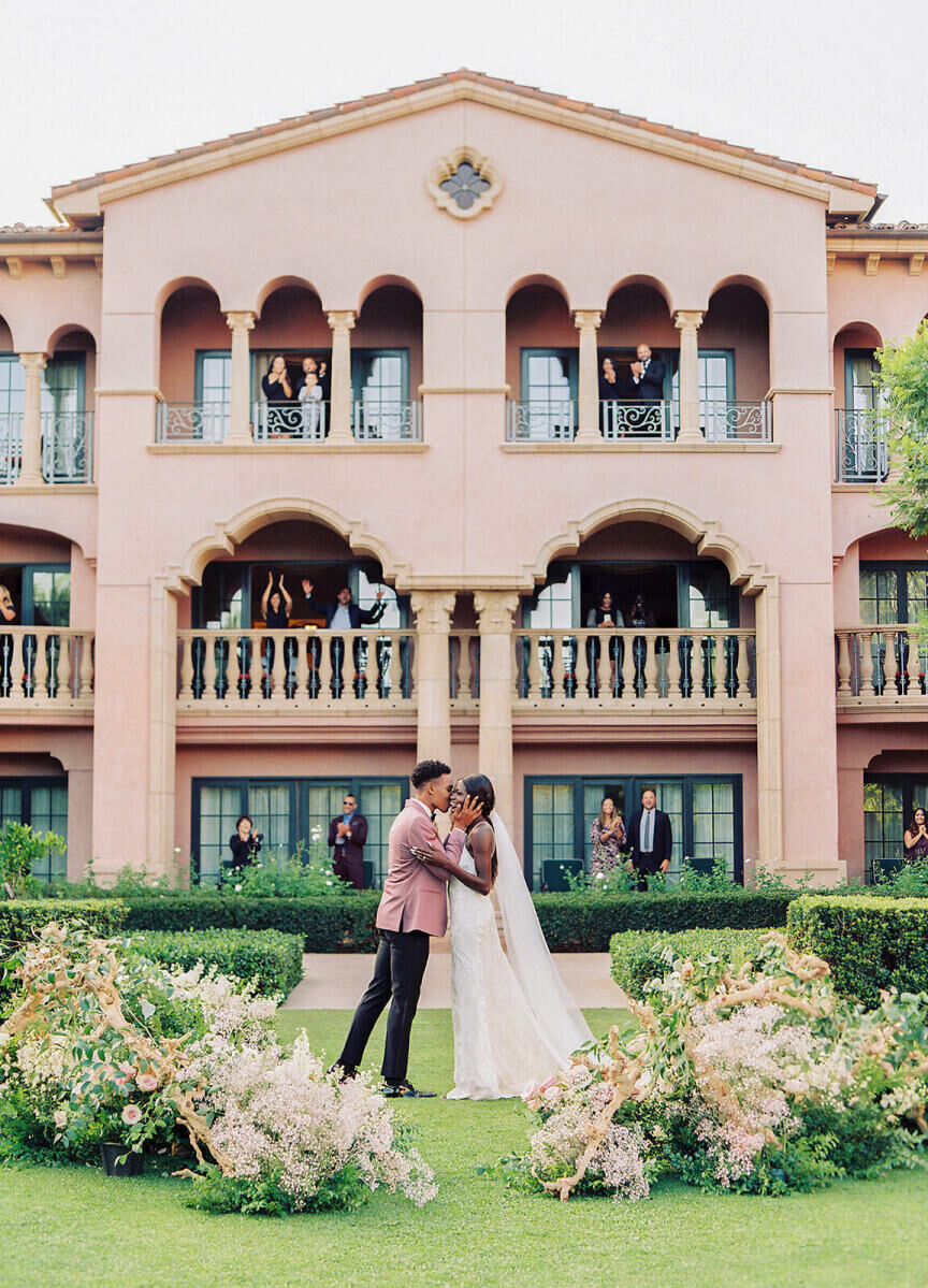 City Weddings: A couple sharing a kiss on their wedding day while guests cheer them on from the pink hotel behind them.