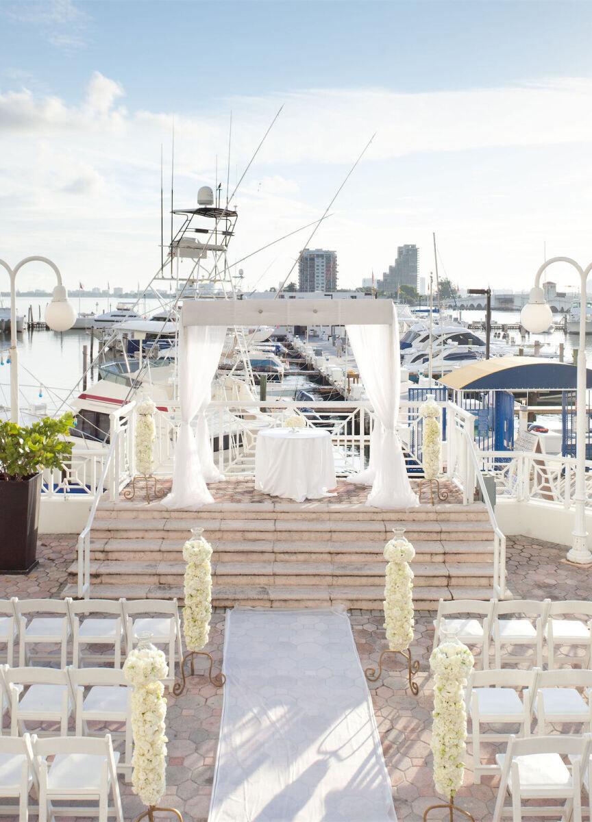 City Weddings: A wedding ceremony setup with white chairs, a white gazebo and white florals, looking over a dock with boats and water beyond it.