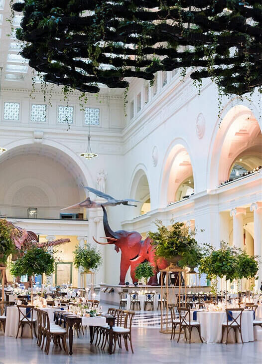 City Weddings: A reception setup at The Field Museum in Chicago, which includes differently sized tables, a hanging greenery installation and elephant.