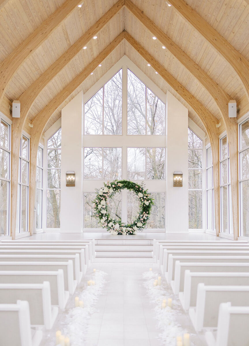 City Weddings: A chapel with a wooden roof, floor-ceiling glass windows, white pews and a circular green arch with white flowers.