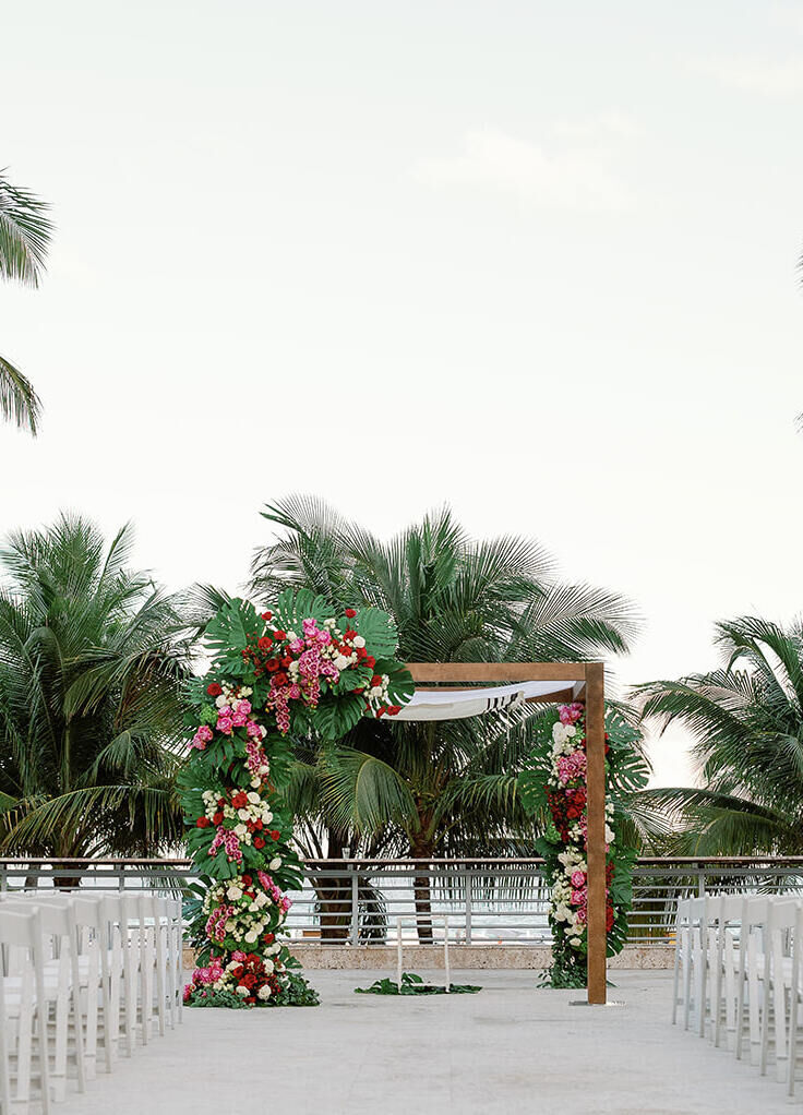 City Weddings: An outdoor wedding reception setup with a wooden arch and white chairs, and palm trees in the background.
