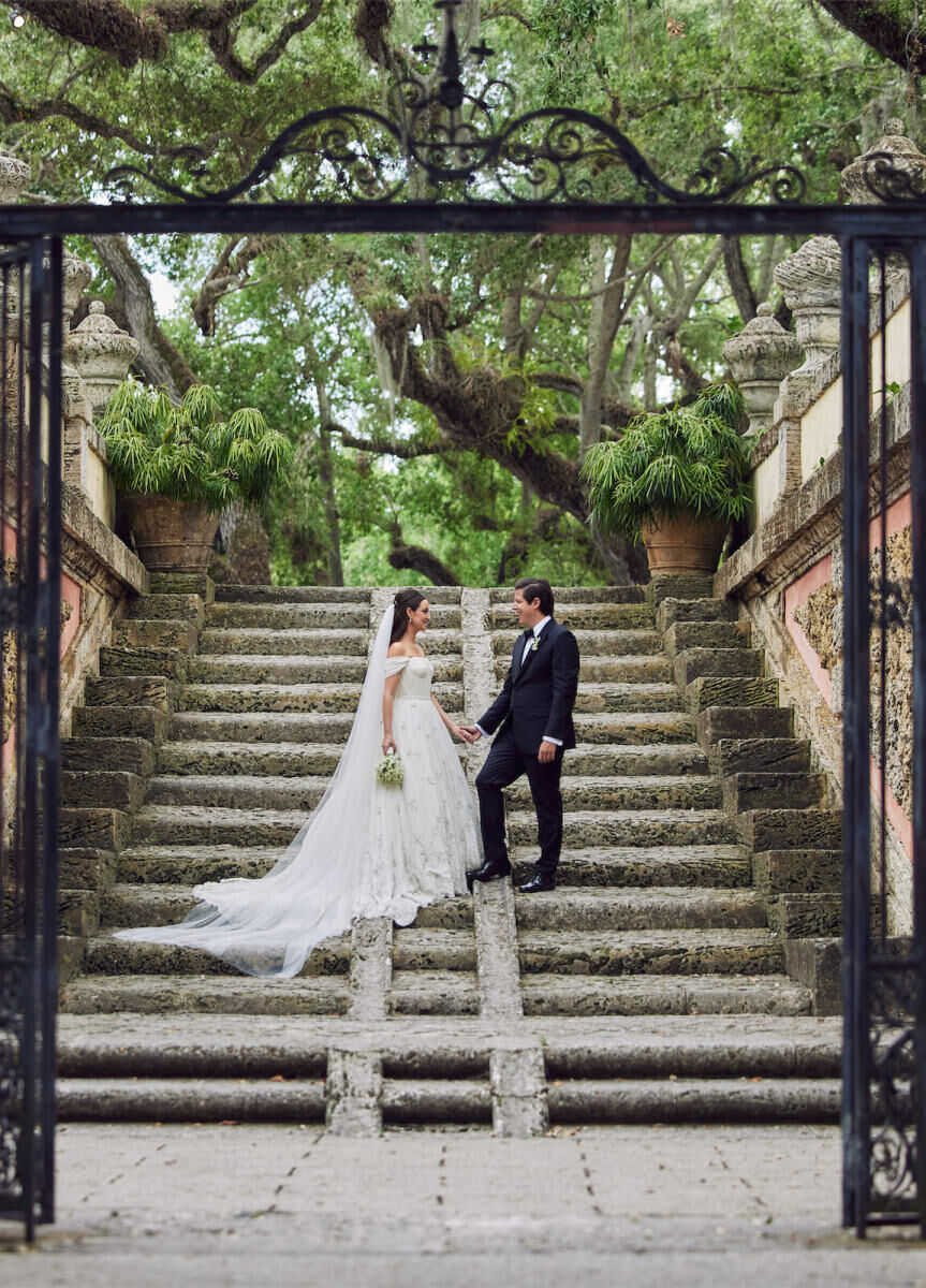 City Weddings: A wedding couple looking at each other and holding hands on a staircase behind an iron gate, with a garden setting beyond them.