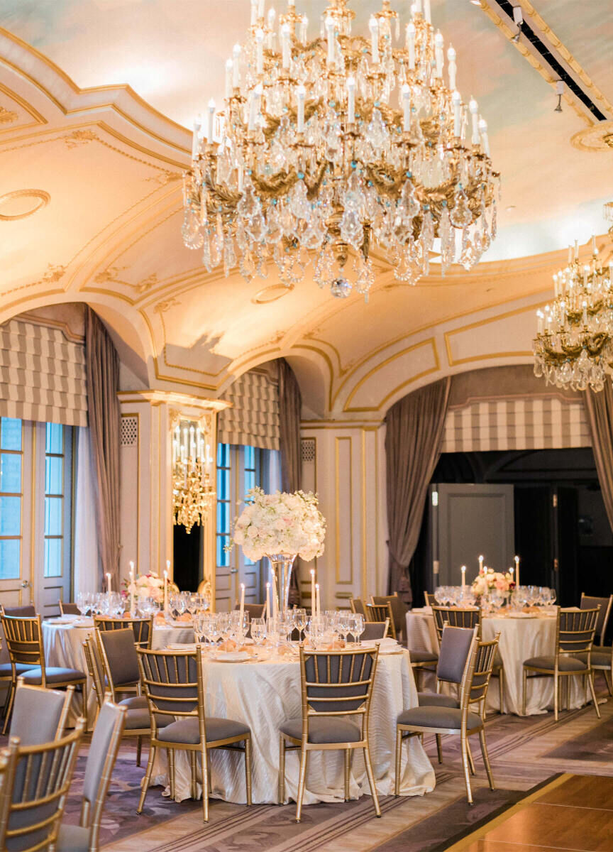 City Weddings: An indoor reception setup in a ballroom with round tables, white floral arrangements in the center and oversize chandeliers above.
