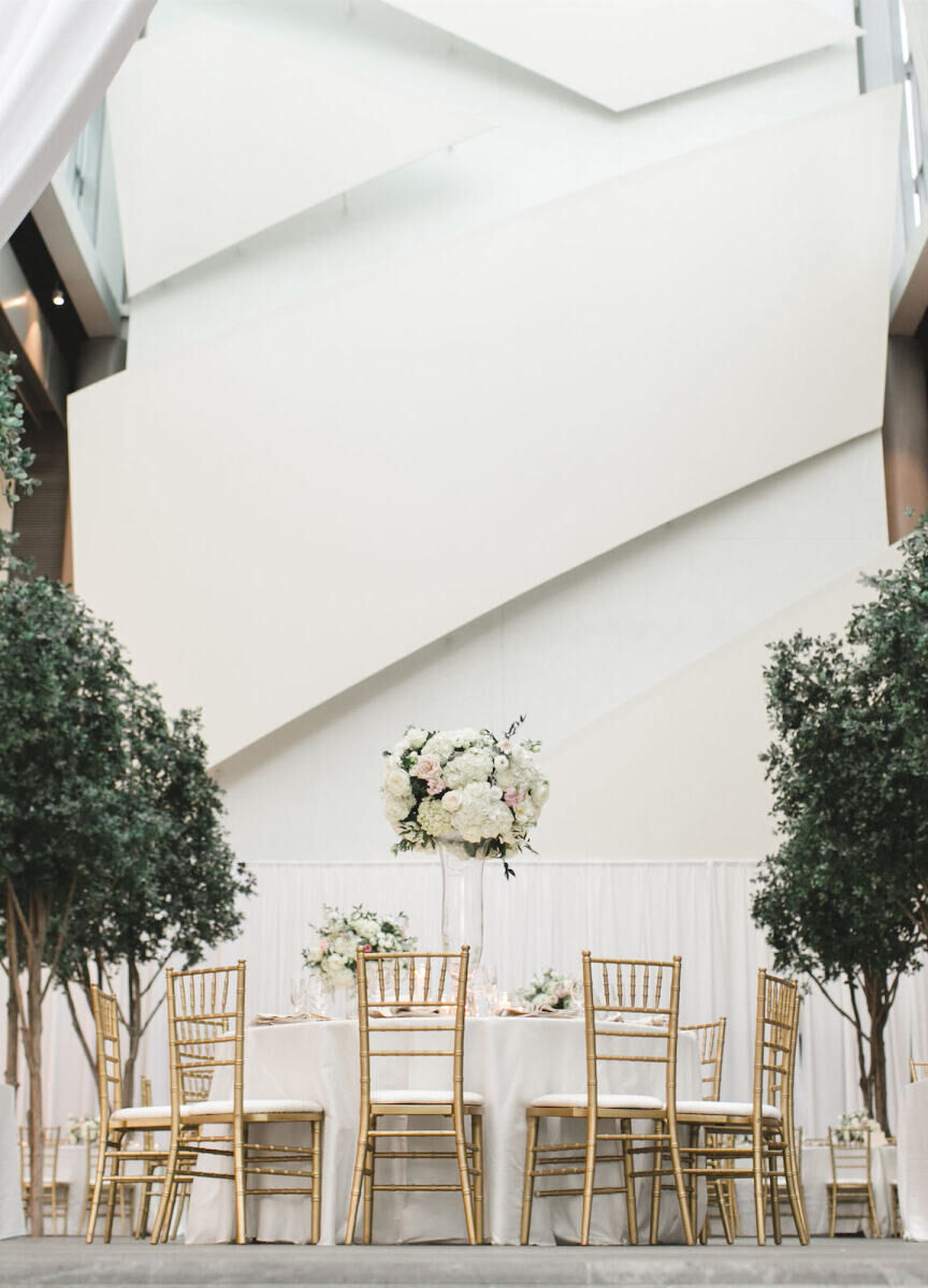 City Weddings: An indoor wedding reception with a white wall, green shrub trees, gold chairs and a round white table with a high white floral arrangement in the center.