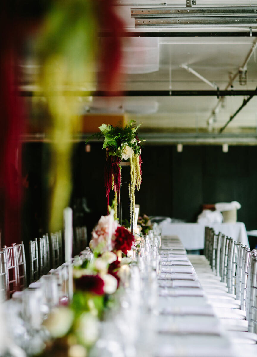 City Weddings: An indoor wedding reception setup with a long rectangular table and tall greenery arrangements at either end.