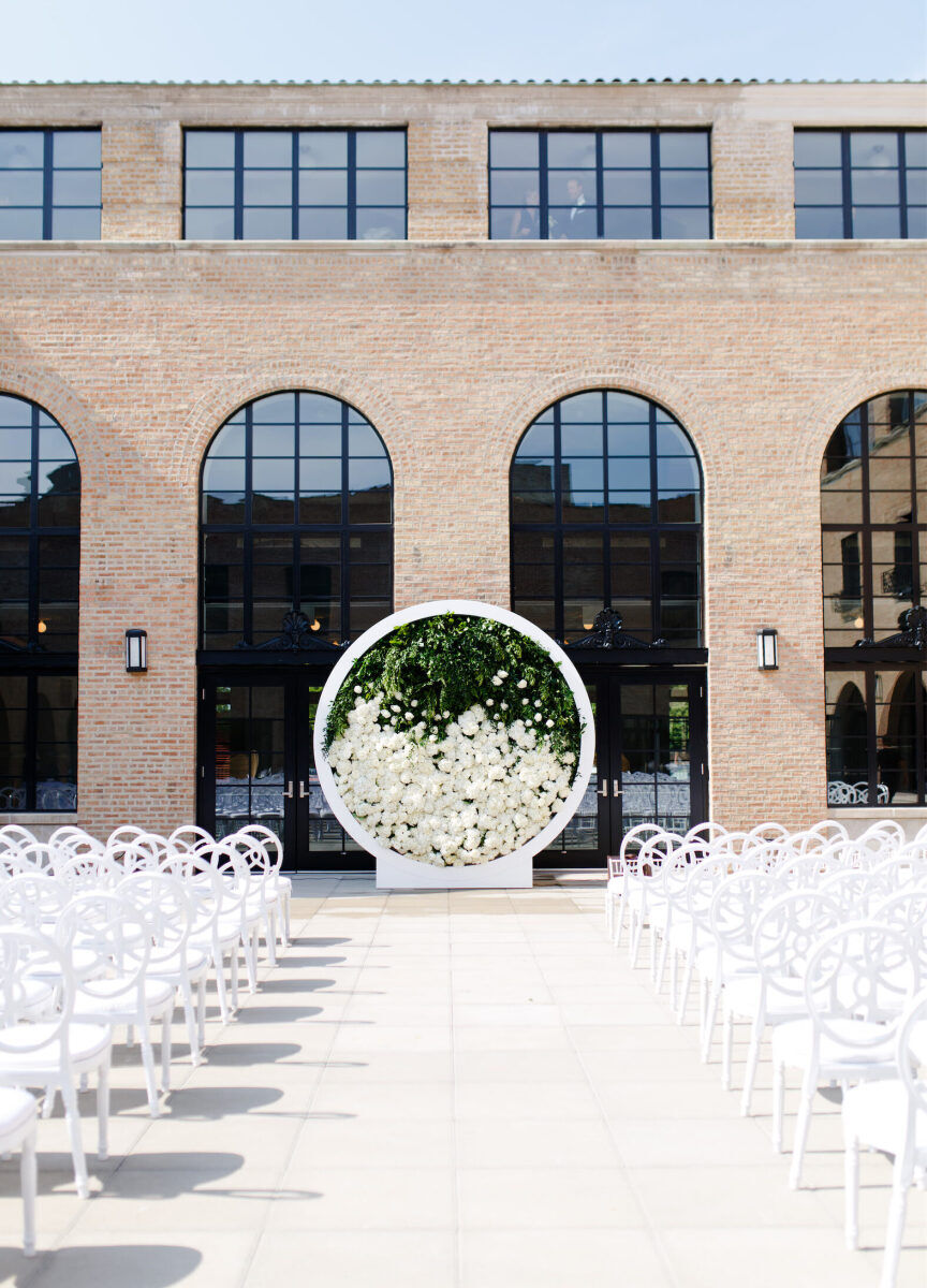 City Weddings: An outdoor wedding ceremony setup with white chairs and a white, circular, flower-filled archway at the end of the aisle.