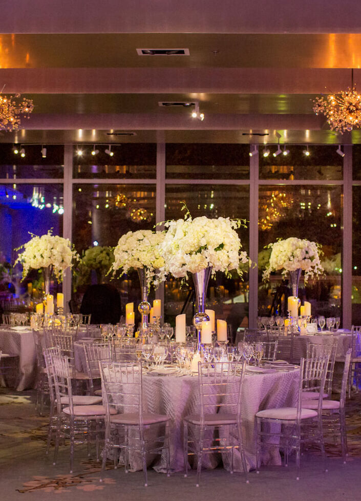 City Weddings: An indoor wedding reception with round tables and high white floral centerpieces by a dance floor.