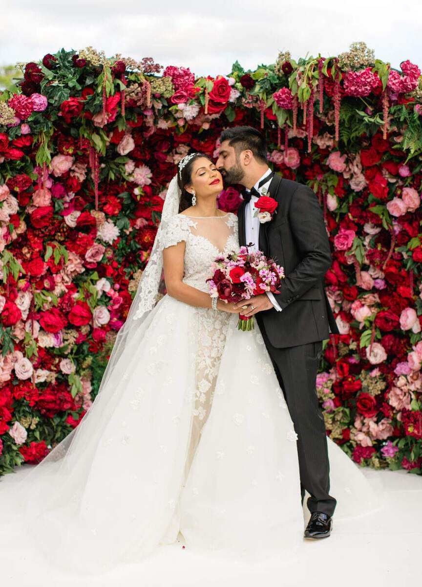 A groom kisses his bride in front of their red and pink floral wall at their colorful countryside wedding.