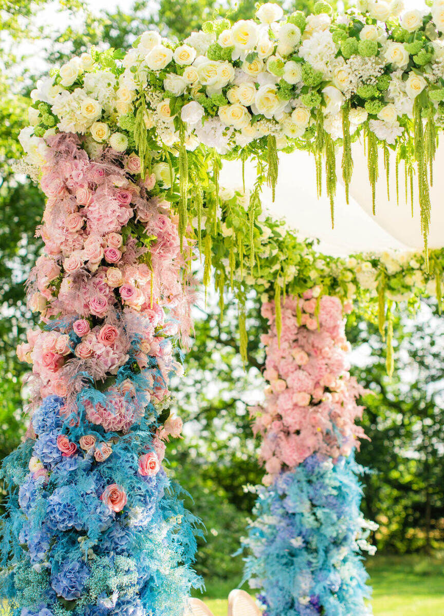A colorful countryside wedding mandap was designed with a gradient of blue and pink flowers and topped with white blooms and dripping greenery.