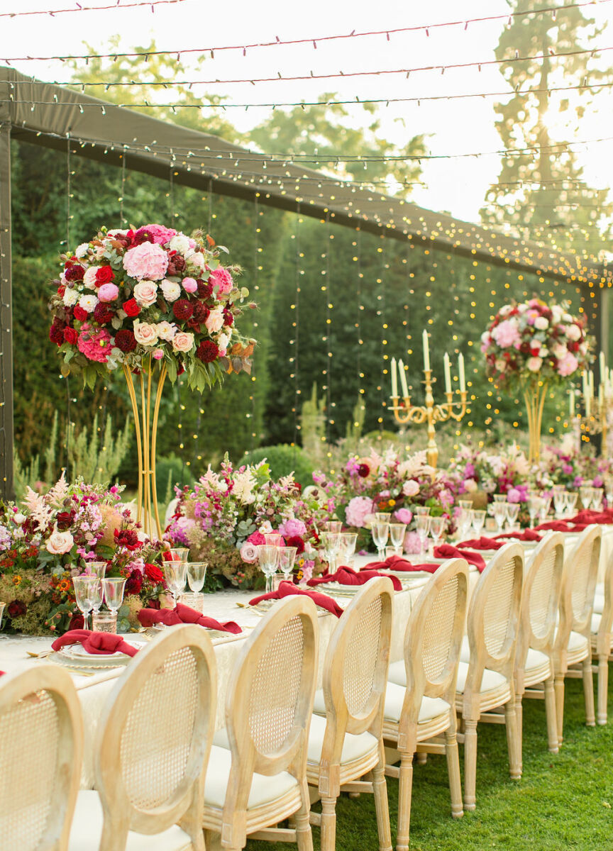 Long tables decorated with high and low centerpieces and red knotted dinner napkins set the scene for a colorful countryside wedding reception outside.