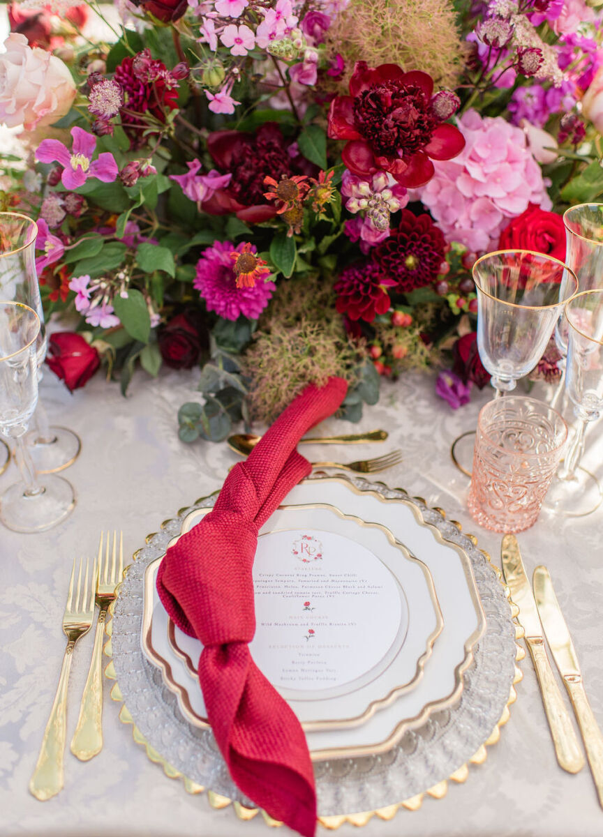 A colorful countryside wedding reception, with a knotted red dinner napkin and pink glassware complementing the lush floral centerpieces.