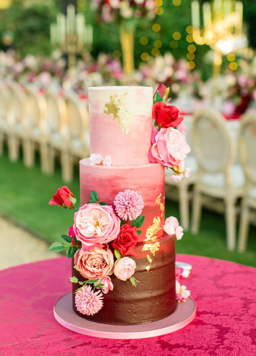 An ombre, three-tier wedding cake in shades of pink and red is decorated by fresh flowers for a colorful countryside wedding reception in England.