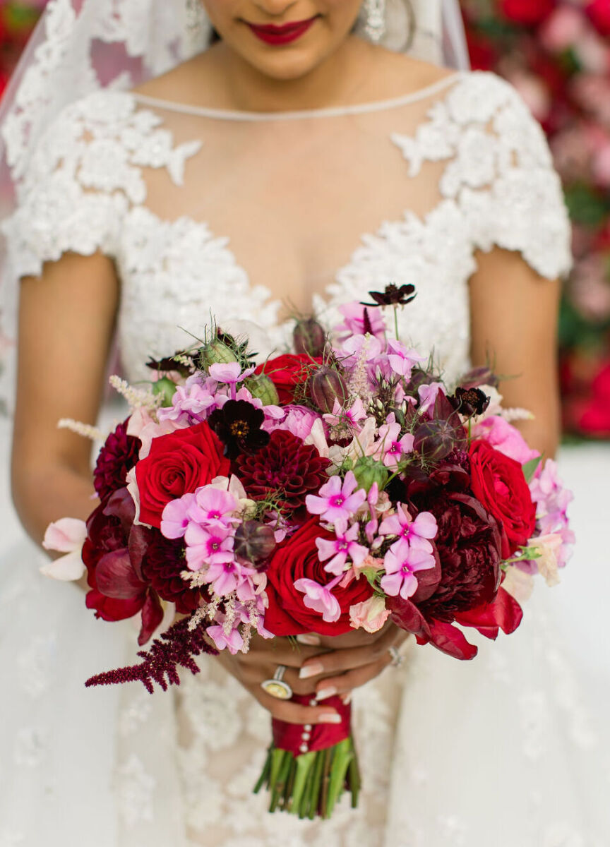 A bride wearing a lace dress and red lipstick holds her bouquet of red and pink flowers at her colorful countryside wedding.