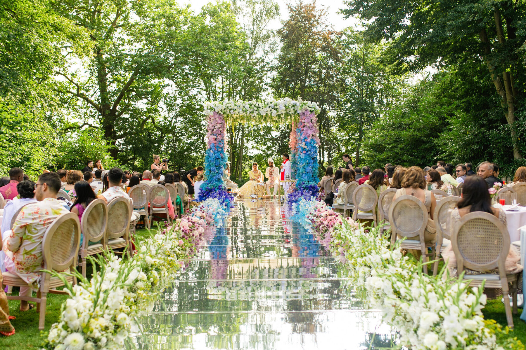 A couple weds under a lush mandap at the end of a mirrored aisle during their colorful countryside wedding ceremony.