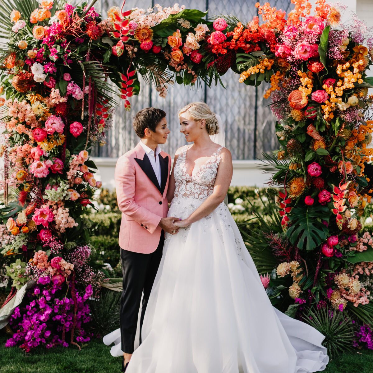 Colorful Wedding: Wedding couple posing beneath a lush ceremony arch of pink and orange blooms at their outdoor wedding ceremony.