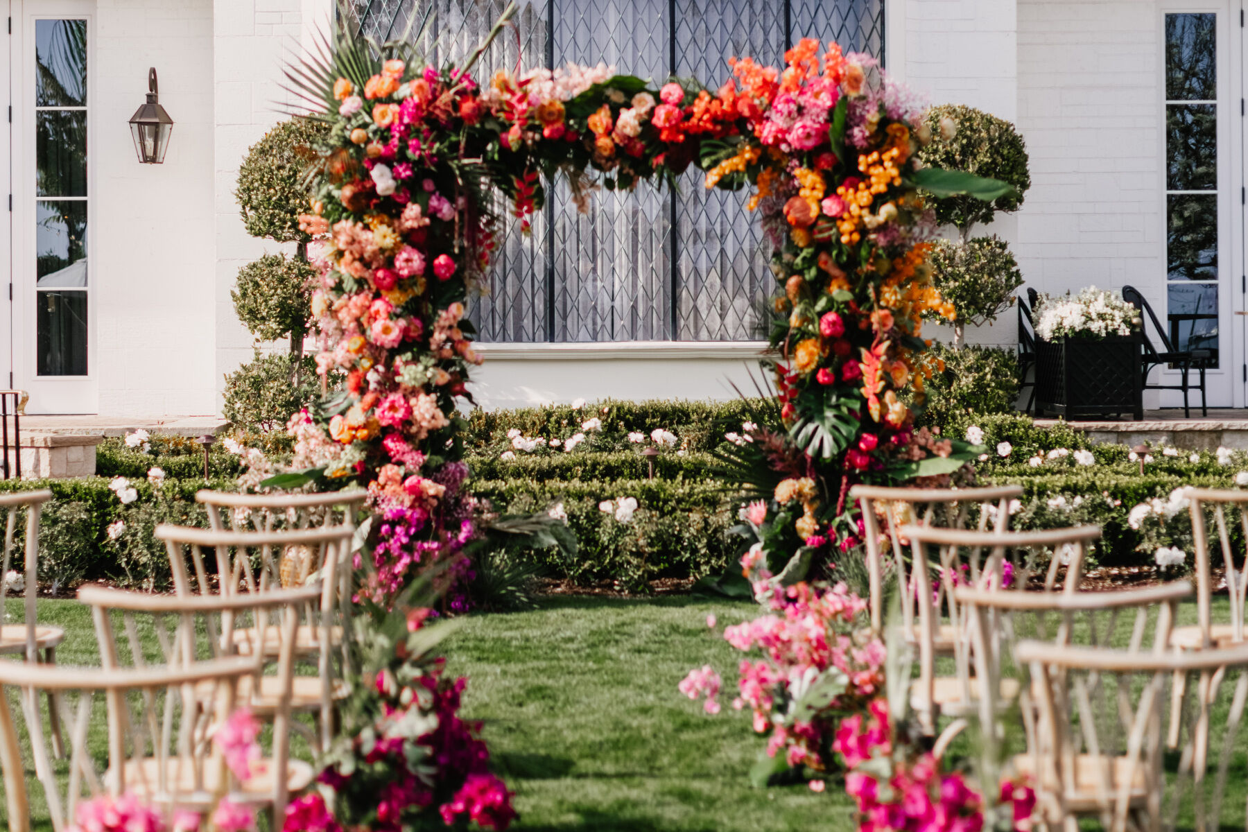 Colorful Wedding: A ceremony setup featuring a floral arch with pink and orange blooms and wooden chairs.