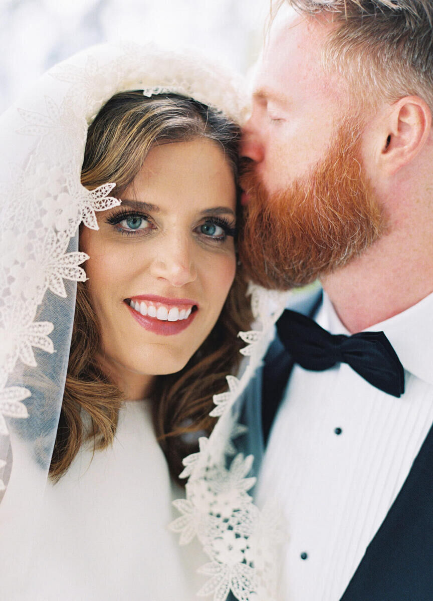 A bride with a retro-glam inspired hair and makeup look, gets a kiss from her husband.