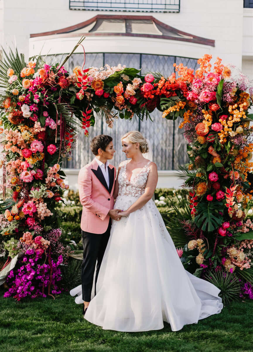 Best Places for a Colorful Wedding: Two newlyweds holding hands and smiling at one another under a colorful floral arch outside at Rosewood Miramar Beach.