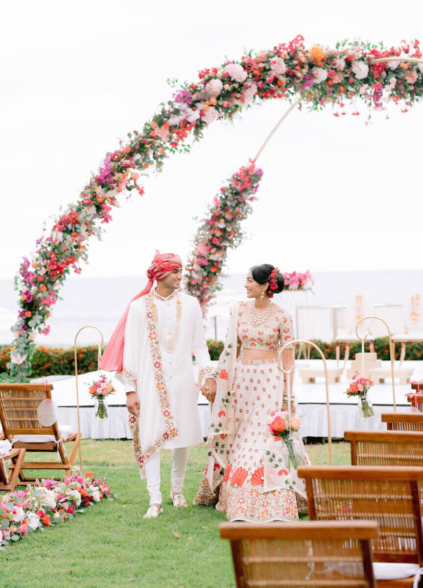 Best Places for a Colorful Wedding: A bride and groom smiling and holding hands walking outside at Grand Velas Riviera Nayarit under a floral arch.