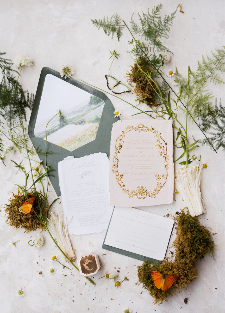A cottagecore wedding invitation suite, with gilded accents and a painted envelope liner depicting the Virginia countryside location, styled with moss, butterflies, mushrooms, and ferns.