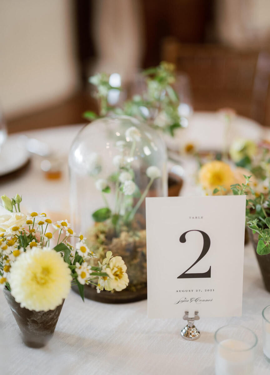 A collection of small centerpieces and simple table number at a cottagecore wedding reception, which incorporated whimsical touches like moss and a mix of fresh and dried flowers.