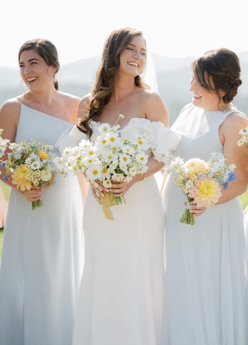 A bride surrounded by her bridesmaids, who wear soft blue gowns and carry bouquets in shades of white, buttery yellow, and blue.