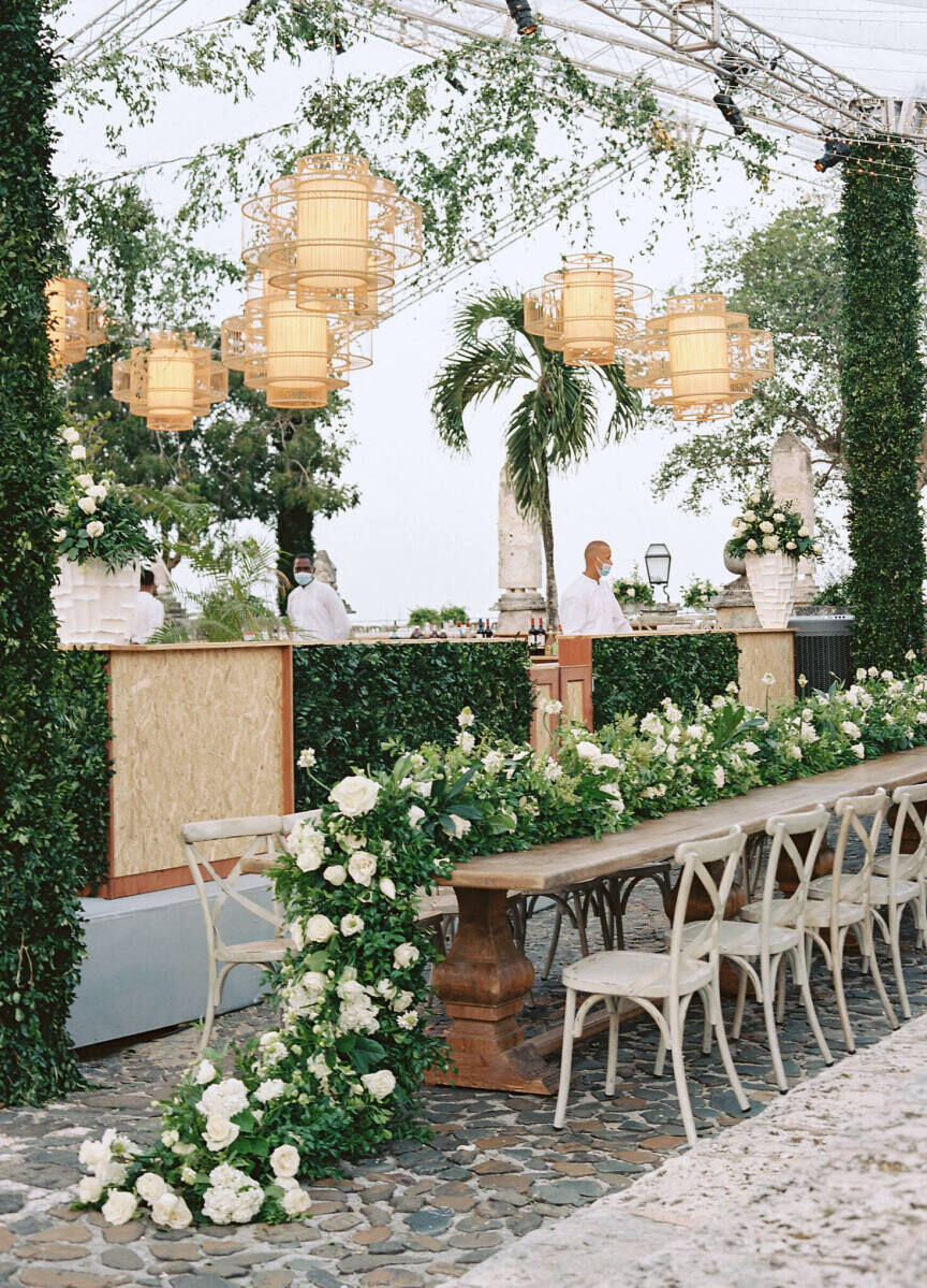 A long table decorated with trailing greenery and white flowers is set in a clear-span tent for a destination wedding reception.
