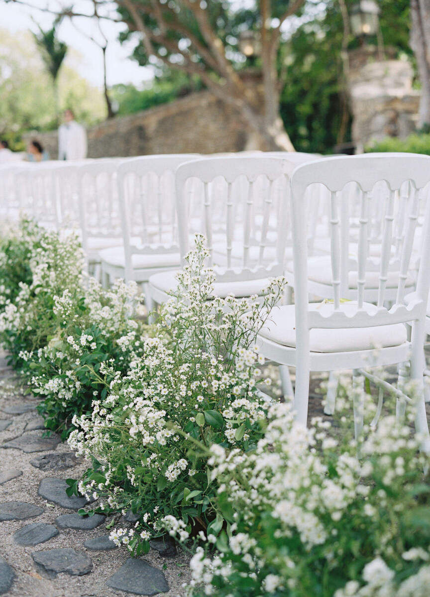 White flowers and greenery line the aisle of this destination wedding ceremony set on a stone plaza.