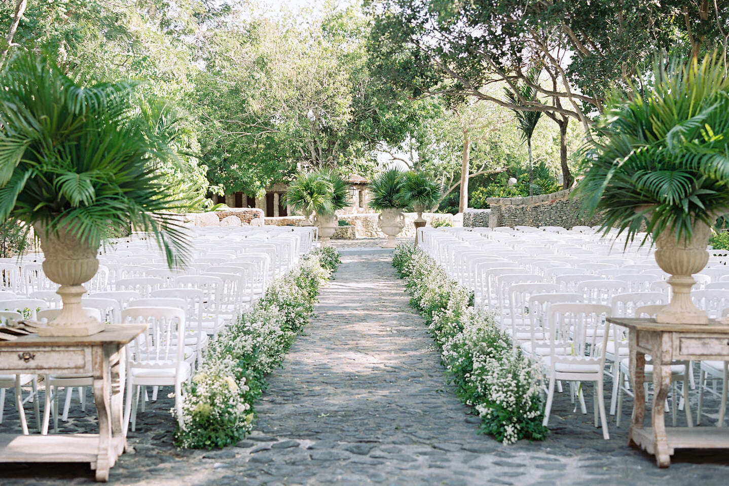 A destination wedding ceremony at a resort in the Dominican Republic, set on a stone courtyard with classic white chairs and white flowers and greenery lining the aisle.